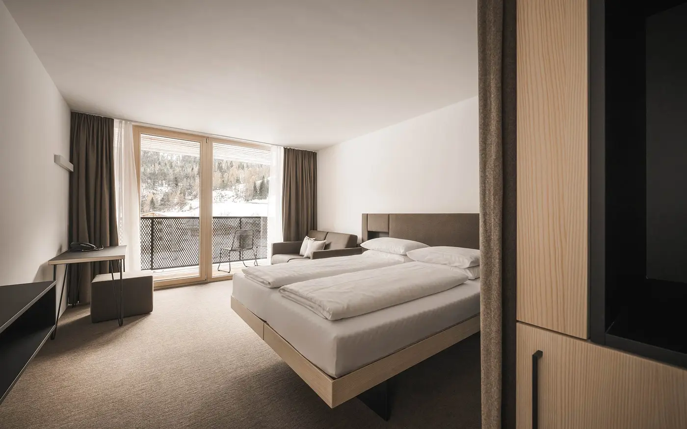 Minimalistisches Hotelzimmer mit Doppelbett, Schreibtisch und großem Fenster mit Blick auf eine verschneite Waldlandschaft.