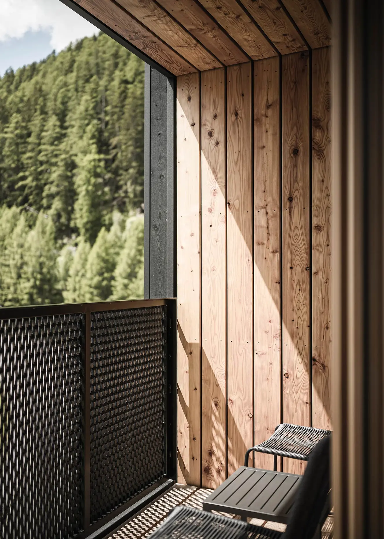 Sonnenbeschienener Holzbalkon mit Metallgeländer und zwei schwarzen Stühlen mit Blick auf einen bewaldeten Hügel.